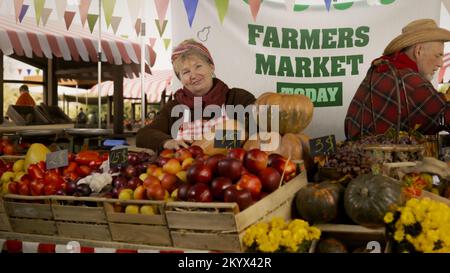 Die Bauernfrau steht am Stand mit frischem, bunten Obst und Gemüse, schaut auf die Kamera. Farmers Market Festival. Vegetarische, biologische und gesunde Lebensmittel. Landwirtschaft. Stockfoto