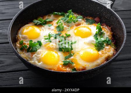Shakshuka-Eier in einer Pfanne vor schwarzer Holzkulisse. Pochierte Eier in einer scharfen Tomaten- und Pfeffersoße. Traditionelles jüdisches Rührei. Oben Stockfoto