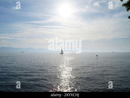 Malerische Landschaft mit einem Segelboot auf dem Bodensee bei Sonnenuntergang auf der Insel Lindau mit den nebligen Schweizer Alpen im Hintergrund an einem Septembertag Stockfoto