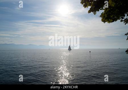 Malerische Landschaft mit einem Segelboot auf dem Bodensee bei Sonnenuntergang auf der Insel Lindau mit den nebligen Schweizer Alpen im Hintergrund an einem Septembertag Stockfoto