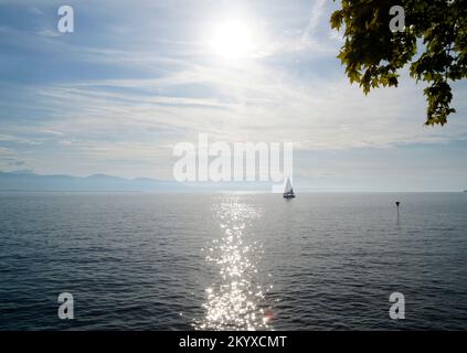 Malerische Landschaft mit einem Segelboot auf dem Bodensee bei Sonnenuntergang auf der Insel Lindau mit den nebligen Schweizer Alpen im Hintergrund an einem Septembertag Stockfoto