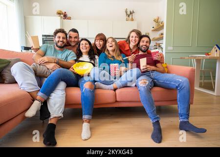 Eine Gruppe von Mitbewohnern schaute fern und aß einen Snack auf der Couch - fröhliche junge Schüler, die Spaß im Wohnzimmer hatten Stockfoto