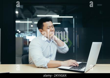 Das Praxisarbeitssyndrom. Ein junger asiatischer Mann sitzt im Büro am Schreibtisch, hält seinen Hals, fühlt Schmerz, Spannung. Er schloss die Augen, massierte mit der Hand. Stockfoto