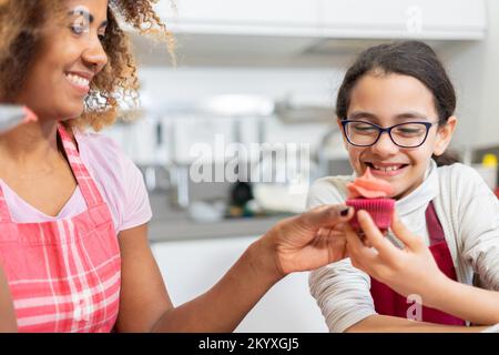 Eine brasilianische Frau, die seiner Tochter zeigt, wie man einen Tassenkuchen dekoriert. Mom und Tochter machen zu Hause zusammen amerikanische Desserts Stockfoto