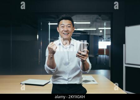 Ein junger, zufriedener asiatischer Geschäftsmann, der ein Spiel am Telefon spielt, hat gute Nachrichten, eine Nachricht. Er steht im Büro am Tisch, schaut in die Kamera, zeigt eine Siegergeste mit seiner Hand. Stockfoto