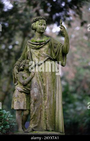 Eine historische Frau mit Kind auf einem alten Friedhof hält ein kleines Kreuz wie ein Selfie-Telefon Stockfoto