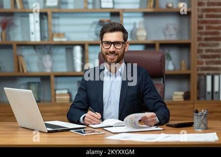 Porträt eines erfolgreichen und glücklichen, ausgereiften Finanziers, eines leitenden Geschäftsmanns mit lächelndem Bart und einer Brille, der in die Kamera schaut, eines Mannes, der im Büro an Papierarbeit arbeitet, eines Investors, der mit dem Ergebnis zufrieden ist. Stockfoto