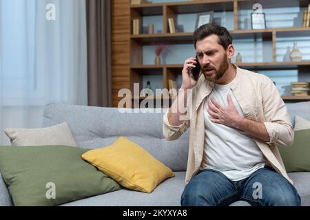 Mann mit Herzinfarkt zu Hause, Patient ruft Arzt am Telefon an, sitzt auf der Couch im Wohnzimmer zu Hause. Stockfoto