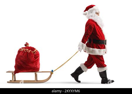 Profilaufnahme über die gesamte Länge, bei der der weihnachtsmann einen hölzernen Schlitten mit einem roten Sack voller Geschenke auf weißem Hintergrund zieht Stockfoto
