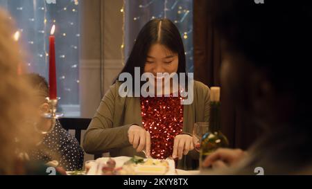 Asian woman eats dish with knife and fork. Multi cultural friends celebrating Christmas or New Year 2023. Served holiday table with candles. Atmosphere of Christmas dinner with friends at home. Stockfoto