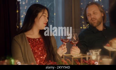 Freunde klingeln, trinken Wein und reden. Multikulturelle Freunde feiern Weihnachten oder Neujahr 2023. Es wurde ein Feiertagstisch serviert. Warme Atmosphäre beim Weihnachtsessen zu Hause. Weihnachtsfeier. Stockfoto