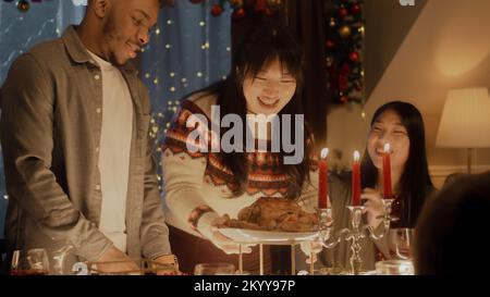 Ein Afroamerikaner schneidet truthahn oder Huhn mit Messer. Multikulturelle Freunde feiern Weihnachten oder Neujahr. Es wird ein Feiertagstisch mit Kerzen serviert. Warme Atmosphäre beim Weihnachtsessen zu Hause. Stockfoto