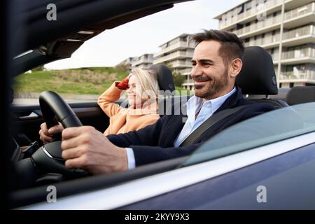 Glückliches Paar mittleren Alters, das mit dem Auto fährt, um sich zu verabreden. Die Frau genießt die Fahrt, während der Ehemann fährt Stockfoto