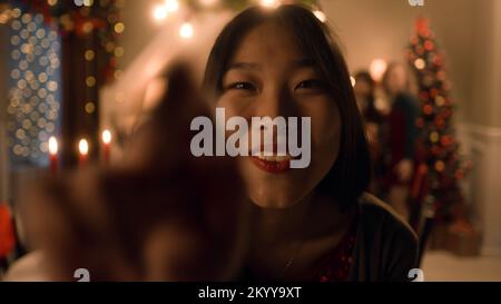 Asiatische Frau stellt den Timer auf Kamera oder Telefon. Glückliche multikulturelle Familie macht Gruppenfotos. Sie feiern Weihnachten oder Neujahr. Tisch mit Geschirr und Kerzen. Weihnachtsessen für die Familie. Kameraansicht. Stockfoto