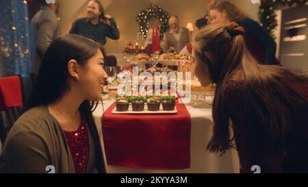 Fröhliche, vielfältige Familie bereitet sich auf das Weihnachtsessen vor: Tisch mit Geschirr decken, Geschenke zeigen, reden und lachen. Asiatische Frau mit Tochter. Warme Atmosphäre im Familienurlaub. Heiligabend Stockfoto