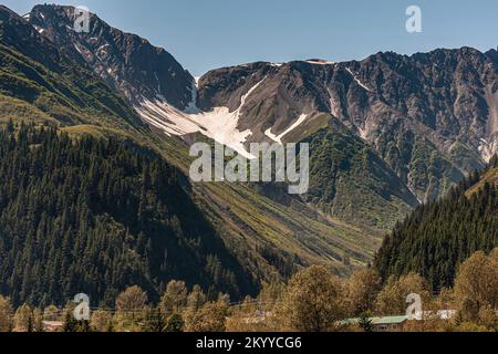 Seward, Alaska, USA - 22. Juli 2011: Landschaft unter blauem Himmel mit dunklen Bergen mit Schneefeltern in Spalten und grünem Wald an den unteren Flanken. T Stockfoto