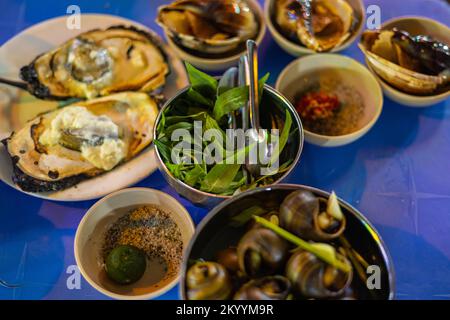 Street Food-Gerichte in den Straßen von Saigon. Muscheln, Schnecken und Austern, zubereitet im lokalen vietnamesischen Stil. Vietnamesische Küche am Abend auf dem Bürgersteig Stockfoto