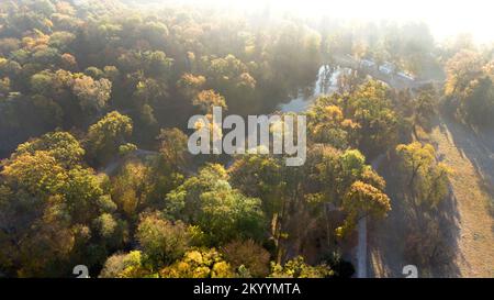 Luftblick über den See mit architektonischen Säulen und Bäumen mit gelb-grünen Blättern im Park an einem hellen, sonnigen Herbstmorgen. Sonnenstrahlen, rotes Sonnenlicht, Sonnenüberstrahlung. Natürlicher Hintergrund Stockfoto