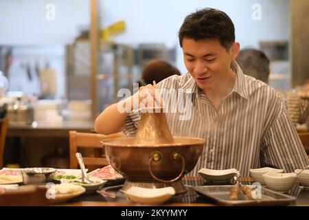 Lächelnder chinesischer junger Mann, der Instant-gekochte Hammel-Hot-Pot im Restaurant isst, ein traditioneller chinesischer Hot-Pot Stockfoto