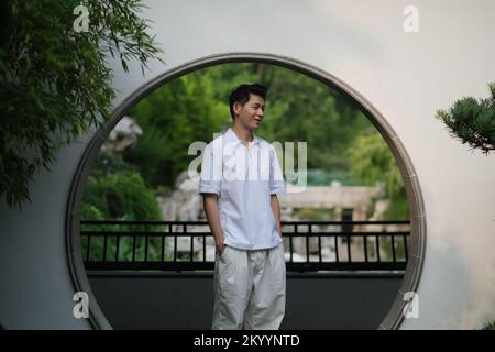 Süßer, lächelnder chinesischer junger Mann am Mondtor im traditionellen chinesischen Garten. Seitenansicht Stockfoto