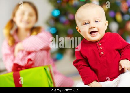 Frohe Weihnachtserinnerungen machen - Babys erstes Weihnachten. Süßer kleiner Junge, der ein Weihnachtsgeschenk mit seiner Schwester im Hintergrund eröffnet. Stockfoto