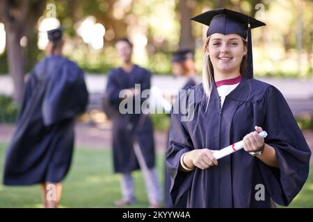 Jetzt kann ich den Job haben, von dem ich immer geträumt habe. Porträt eines lächelnden Absolventen, der sein Diplom hält, mit ihren früheren Absolventen im Hintergrund. Stockfoto