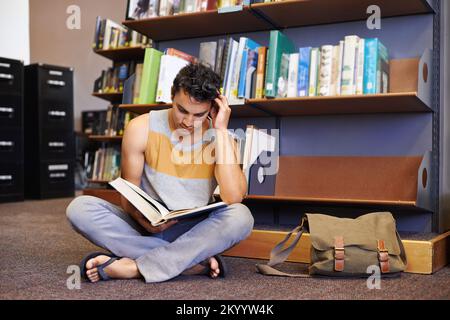 Er lernt wirklich hart für das kommende Finale. Junger Student, der auf dem Boden einer Bibliotheksleserin sitzt. Stockfoto