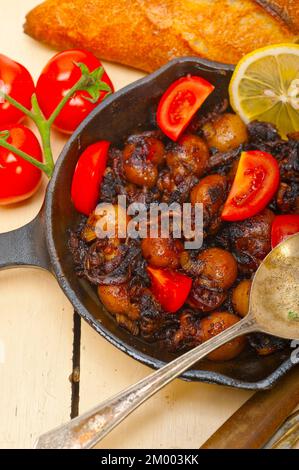 Baby Tintenfisch über rustikalen Holztisch auf eisernen Pfanne mit Tomaten und Zwiebeln gebraten Stockfoto