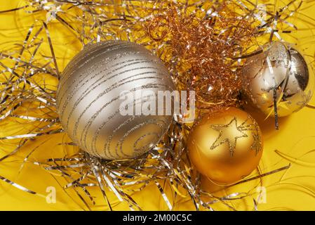 Drei dekorative silberne und goldene weihnachtskugeln auf einem Bündel metallischer Fäden auf gelbem Hintergrund. Stockfoto