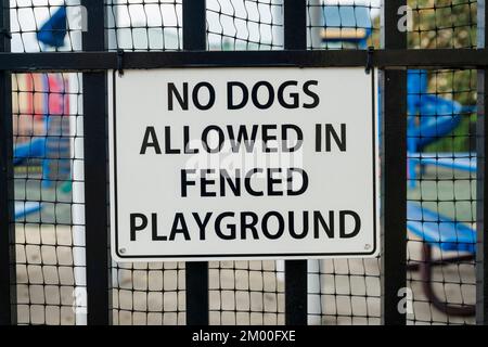 Nahaufnahme einer Beschilderung, bei der Hunde auf dem eingezäunten Spielplatz nicht erlaubt sind. Unterschreiben Sie auf einem bemalten schwarzen Geländer mit Metallschirm außerhalb des Spielplatzes. Stockfoto