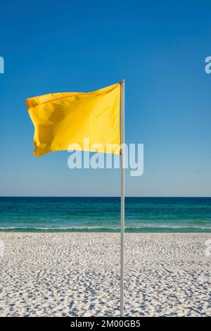 Gelbe Flagge an einem Stab am Strand in Destin, Florida. Vertikaler Schuss einer Flagge mit Blick auf einen weißen Sand und blauen Ozean und Himmel am Hintergrund Stockfoto