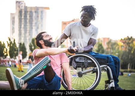 Zwei Freunde mit unterschiedlichen körperlichen Behinderungen begrüßen einander im Park, ein erwachsener afrikanischer Mann im Rollstuhl, der sich mit seinem hispanischen freitag die Hand schüttelt Stockfoto