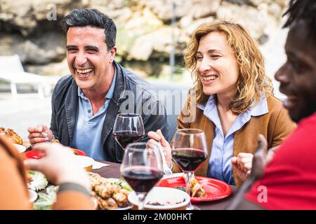 Erwachsene, die beim Grillen essen und Spaß haben, Rotwein zusammen auf einer Familienfeier zu trinken, glückliche Freunde, die grillspezialitäten im Restaurant im Freien essen, echt Stockfoto