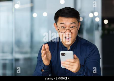 Nahaufnahme eines Mannes mit Brille und Hemd, schockiert vom Lächeln der guten Nachricht und dem Blick auf den Laptop-Bildschirm, erfolgreicher asiatischer Geschäftsmann, der in einem modernen Bürogebäude arbeitet. Stockfoto