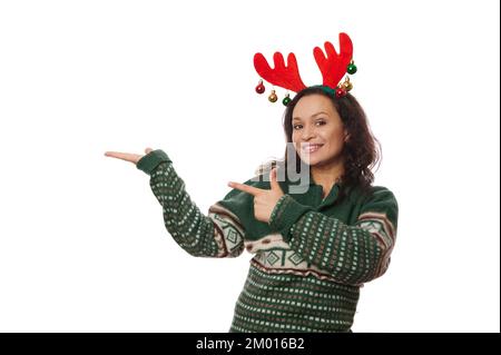 Fröhliche Brünette in weihnachtsgrünem Pullover und Hirschgeweih-Korb, hält und zeigt auf Werbetexte auf der Handfläche nach oben Stockfoto