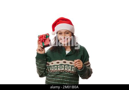 „Overjoyed Pretty Woman“ mit Weihnachtsmannmütze und grünem Pullover mit Weihnachtsgemusterung, hält ein Weihnachtsgeschenk isoliert auf weißem Hintergrund Stockfoto