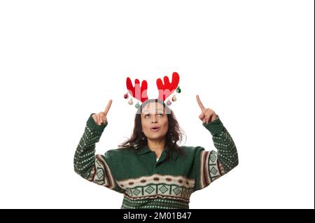 Fröhliche Brünette in Xmas grünem Pullover und Hirschgeweih-Korb, der auf das Kopierfeld zeigt, über ihrem Kopf auf weißem Hintergrund Stockfoto
