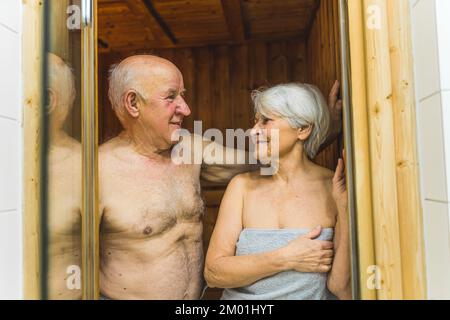 Europäisches weißes, heterosexuelles Ehepaar, das sich gegenseitig in die Augen sieht, lächelt und ein SPA-Erlebnis genießt. Die Ehe sieht man durch die gläserne Saunatür. Indoor-Aufnahme. Hochwertiges Foto Stockfoto