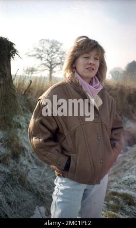 1980er, historisch, draußen an einem frostigen Morgen, mit klarem Himmel, stand eine junge Frau Mitte zwanzig, die eine braune Lederjacke der Epoche, eine Bomberjacke mit breiten Schultern, einen rosa Schal und verblasste hellblaue Jeans trug, England, Großbritannien. Bomber- oder Pilotenjacken waren in den 1980er Jahren beliebte Modeartikel, insbesondere nach der Veröffentlichung des erfolgreichen Films Top Gun im Jahr 1986. Stockfoto