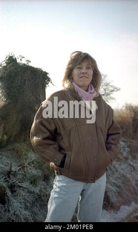 1980er, historisch, draußen an einem frostigen Morgen, mit klarem Himmel, stand eine junge Frau Mitte zwanzig, die eine braune Lederjacke der Epoche, eine Bomberjacke mit breiten Schultern, einen rosa Schal und verblasste hellblaue Jeans trug, England, Großbritannien. Bomber- oder Pilotenjacken waren in den 1980er Jahren beliebte Modeartikel, insbesondere nach der Veröffentlichung des erfolgreichen Films Top Gun im Jahr 1986. Stockfoto