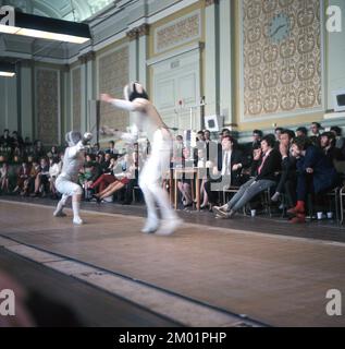 1970er, historischer Zäunwettbewerb, zwei Fechter, die auf einer langen Matte gegeneinander kämpfen, in einem großen Saal vor einer Gruppe sitzender Zuschauer, England, Großbritannien. Stockfoto