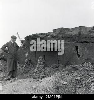 1950er, historisch, auf der Baustelle eines Stahlwerks, stand ein Arbeiter in Overalls und flacher Kappe und schaute auf eine alte Betonstruktur, möglicherweise die Überreste eines alten Bunkers aus dem Jahr WW1, nahe Swansea Bay in Port Talbot, South Wales, Großbritannien. Stockfoto