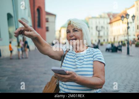 Eine Hand hält ein Handy mit einem leeren Bildschirm, der auf eine Straße in der Stadt zeigt. 50s 60s Frau in lässiger Kleidung, die ein Mobiltelefon benutzt, zeigt das Stockfoto