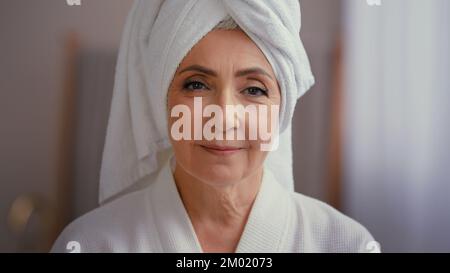 Alte kaukasische Frau mittleren Alters, ältere Dame, weibliches Modell mit Handtuch auf dem Kopf im Bademantel, die auf die Kamera schaut und Geste der Stille zeigt Stockfoto