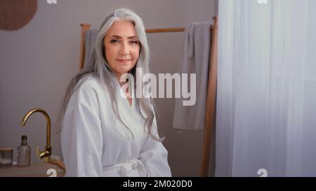 Porträt wunderschöne 60s alte, weiße 50s ältere, reife Frau, die im Badezimmer steht. Großmutter, weibliche Frau mit grauen Haaren, die Bademantel trägt und drinnen posiert Stockfoto