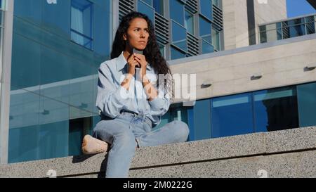 Eine pensive gestresste, traurige Frau mit Handy sitzt in der Stadt in der Nähe eines Gebäudes und denkt über Probleme nach, um nach Lösungen zu suchen Sorgen über niedrigen Akkustand Stockfoto