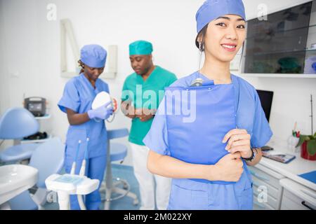 Asiatische Zahnärztin in einer Zahnarztpraxis mit ihren Kollegen, die hinter ihrer multiethnischen Gruppe sprechen Stockfoto