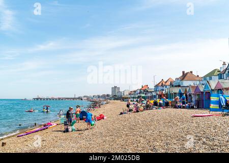 Herne Bay Beach an einem heißen Sommertag. Strandhütten bemalten verschiedene Farben hintereinander mit Menschen am steinigen Strand unter blauem Himmel. Pier in der Ferne. Stockfoto