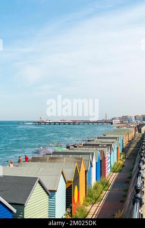 Herne Bay am Meer an einem heißen Sommertag. Eine Reihe von Strandhütten mit dem geschäftigen Strand, dem Pier und dem Uhrenturm in der Ferne unter blauem Himmel. Stockfoto