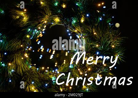 Frohe Weihnachten Text auf immergrünen Winterzweigen, die von Schnee mit Bokeh-Lichtern bedeckt sind. Stockfoto